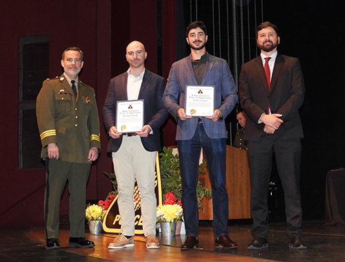 Four men stand on a stage; two are holding certificates.